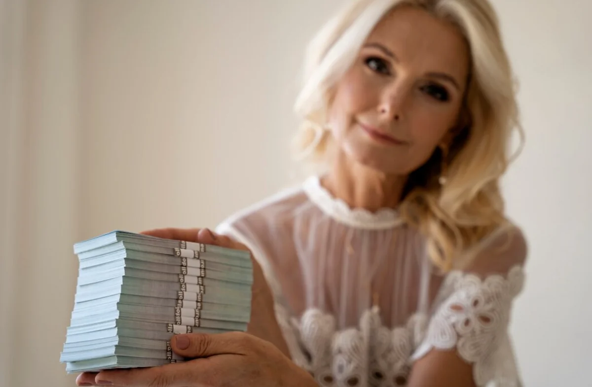 Blond kobieta w białej bluzce trzyma w dłoniach duży stos banknotów.
