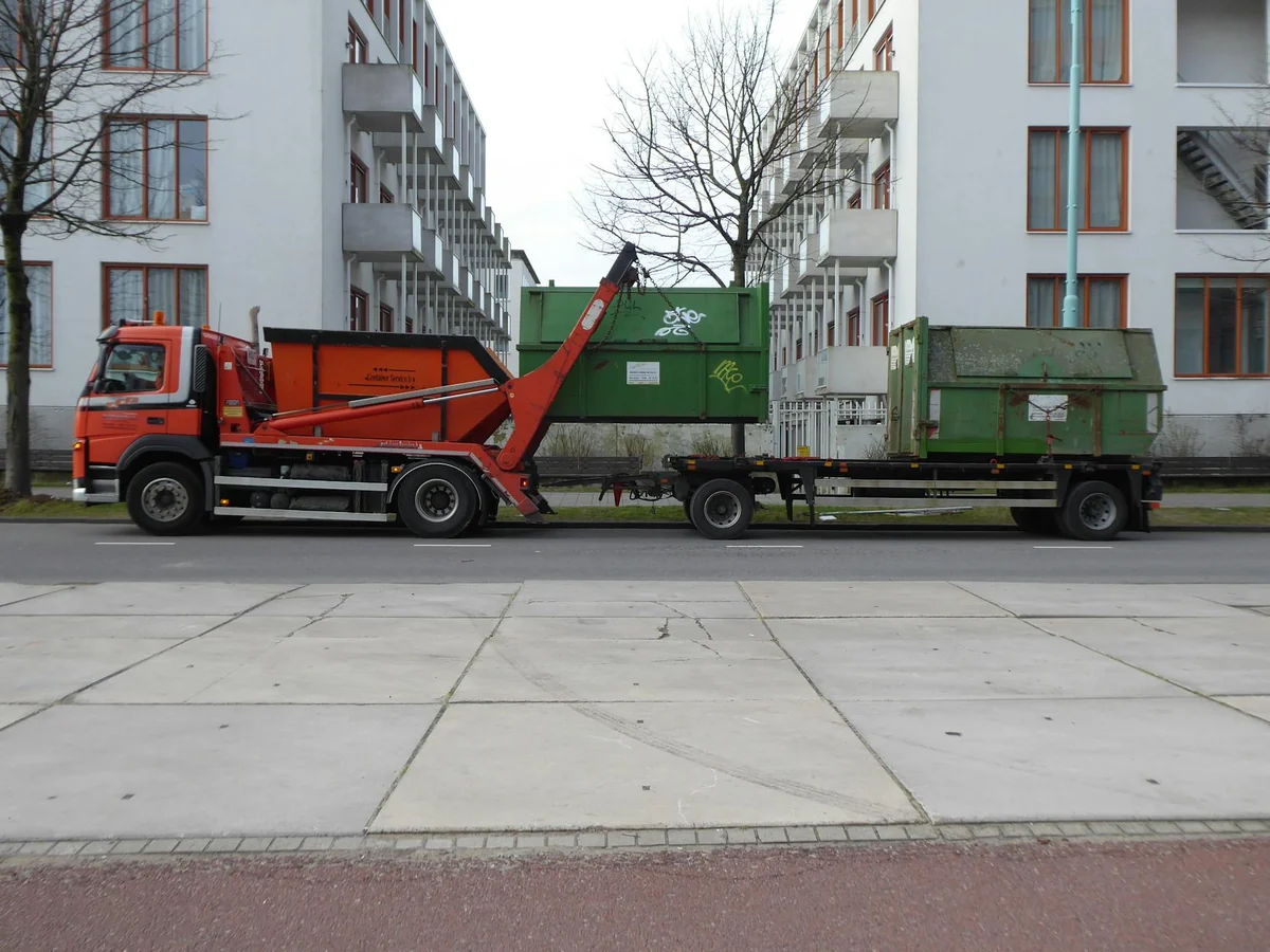 Pomarańczowa ciężarówka z kontenerami na tle bloków mieszkalnych.