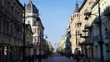 Deptak z zabytkowymi kamienicami, ozdobnymi latarniami i przechodniami; w oddali pomnik.