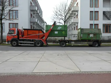Pomarańczowa ciężarówka z kontenerami na tle bloków mieszkalnych.