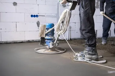 Robotnicy zacierają świeżą posadzkę betonową maszyną w niedokończonym wnętrzu.