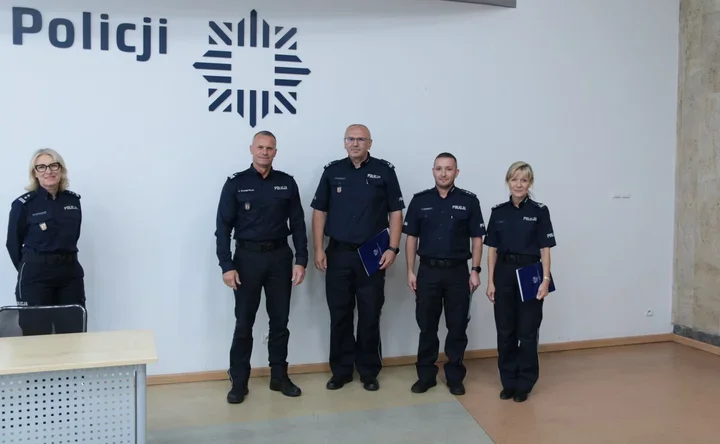 Pięcioro funkcjonariuszy policji w mundurach stoi przed białą ścianą z napisem „Policji” i logo, niektórzy trzymają niebieskie teczki.
