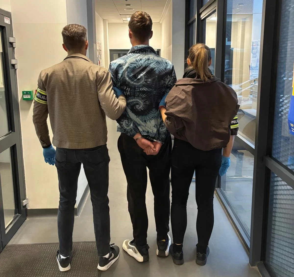 Funkcjonariusze policji prowadzą zatrzymanego mężczyznę z rękoma skutymi kajdankami korytarzem budynku.