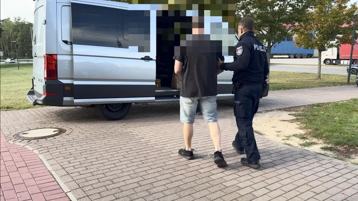 Funkcjonariusz policji prowadzi mężczyznę w stronę srebrnego furgonu stojącego na brukowanej nawierzchni przy drodze.