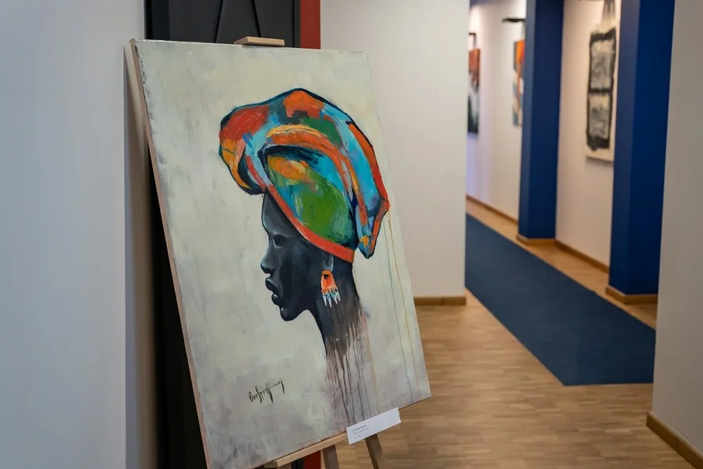 W galerii na sztalugach stoi obraz olejny przedstawiający profil kobiety w kolorowym turbanie, w tle korytarz z wystawionymi pracami.