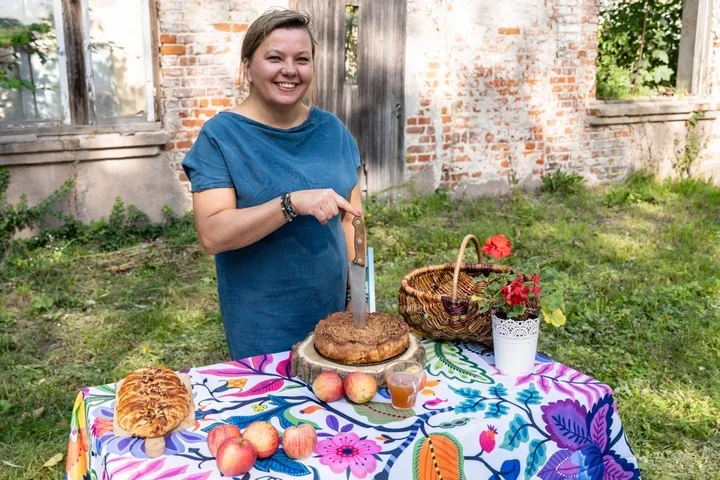 Kobieta w niebieskiej sukience kroi nóżem jabłecznik na kolorowym stole w ogrodzie przed starą ceglaną kamienicą.