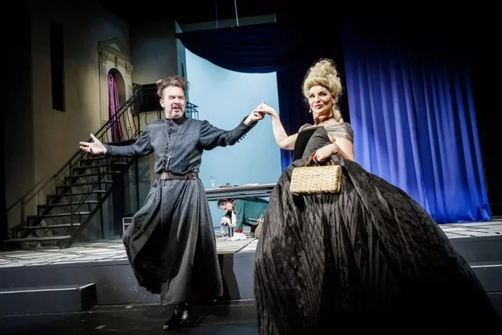 Aktor w kostiumie duchownego i aktorka w czarnej sukni z krynoliną tańczą na scenie teatralnej.