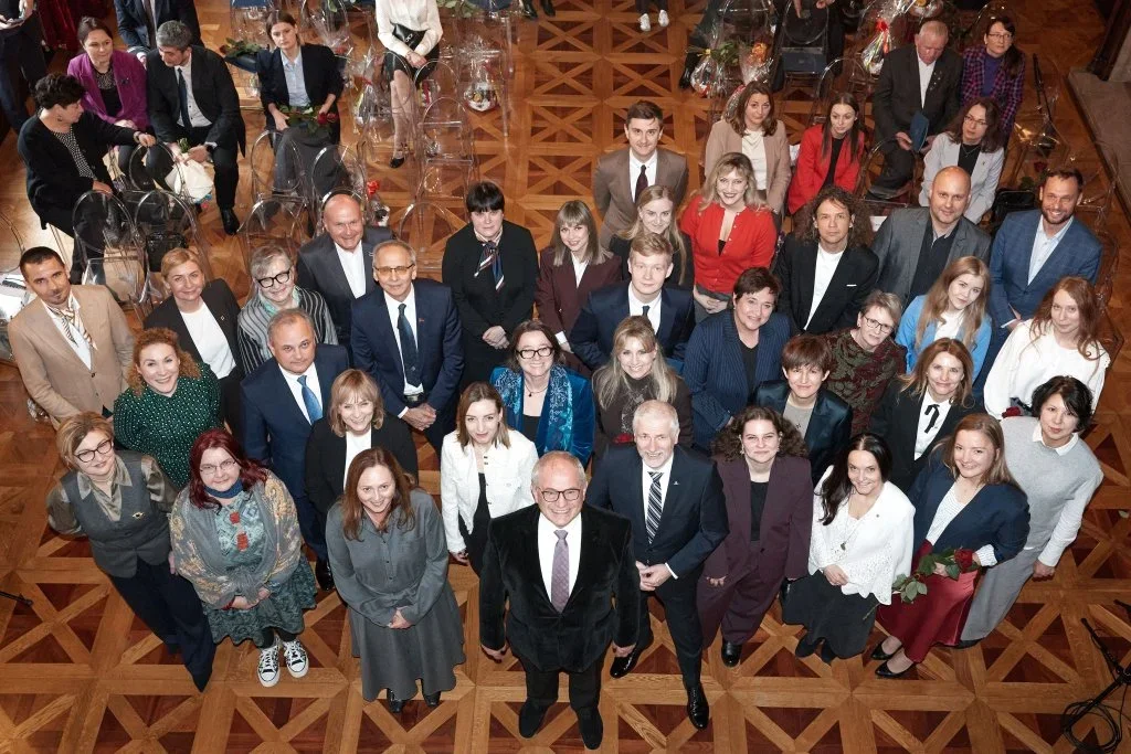 Grupa osób pozuje do pamiątkowego zdjęcia grupowego w reprezentacyjnej sali z drewnianą parkietową podłogą.