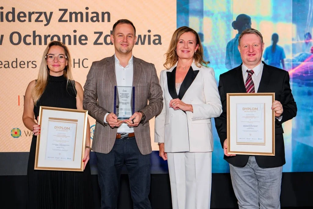 Cztery osoby stoją na scenie podczas uroczystości wręczenia nagród w ochronie zdrowia, dwie z nich trzymają dyplomy, a jedna statuetkę.