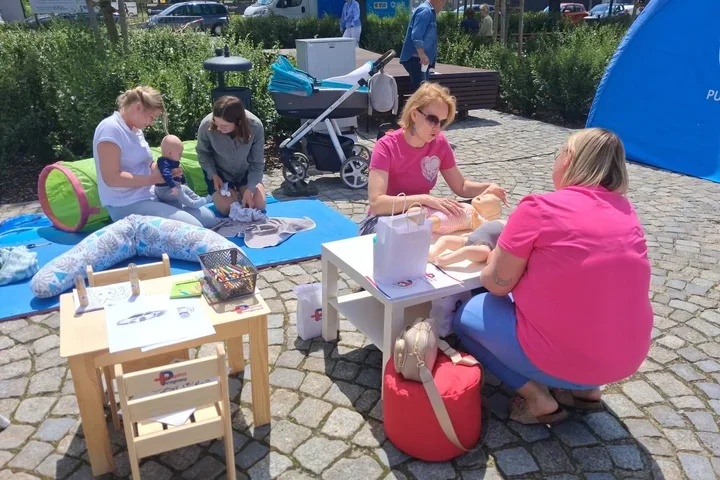 Kobiety z dziećmi biorą udział w plenerowej akcji edukacyjnej dotyczącej pierwszej pomocy dla niemowląt, na brukowanym placu obok niebieskiego namiotu.