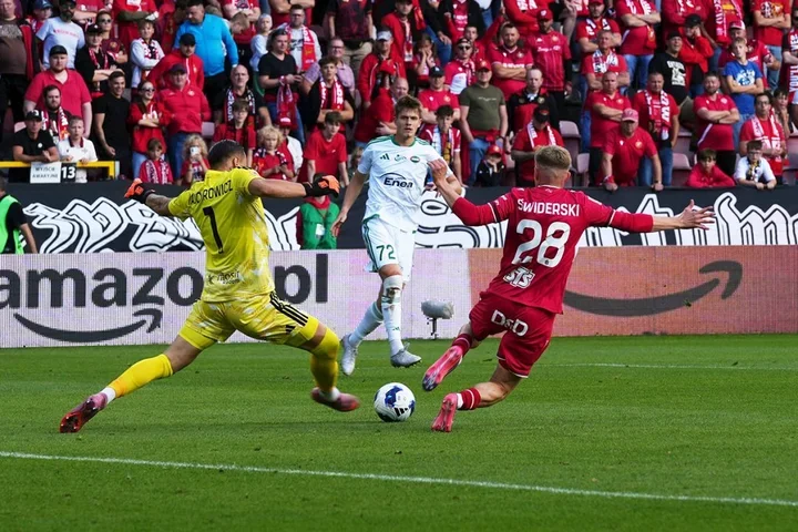 Piłkarz w czerwonym stroju o nazwisku Świderski strzela do bramki, a bramkarz w żółtym stroju o nazwisku Majchrowicz próbuje obronić podczas meczu na stadionie