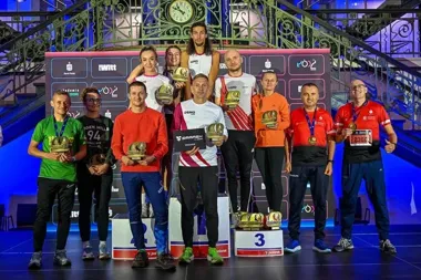 Grupa biegaczy stoi na podium z trofeami podczas ceremonii wręczenia nagród w hali z ozdobną klatką schodową.