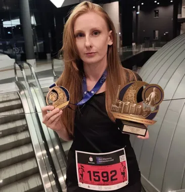 Uczestniczka biegu PKO Wieczorny Półmaraton Łódź z numerem 1592 stoi w budynku obok ruchomych schodów i trzyma medal oraz pamiątkową statuetkę z datą 19.09.2026