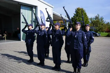 Kobiety w granatowych mundurach i czapkach stoją w szyku na brukowanym placu, trzymając karabiny uniesione w górę, podczas uroczystości przed budynkiem.