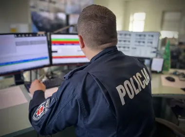 Policjant w mundurze siedzi przed monitorami w centrum dowodzenia lub na stanowisku dyżurnym.