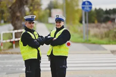 Dwóch policjantów w odblaskowych kamizelkach i czapkach stoi z założonymi rękami na przejściu dla pieszych przy drodze.
