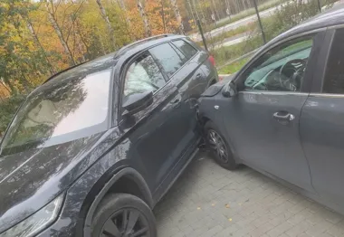 Dwa samochody osobowe – czarny SUV i szary sedan – stykają się bokami na brukowanym parkingu w otoczeniu drzew z jesiennym liściem.