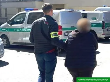 Funkcjonariusz Straży Granicznej prowadzi zatrzymaną osobę przed pojazdami służbowymi na ulicy.