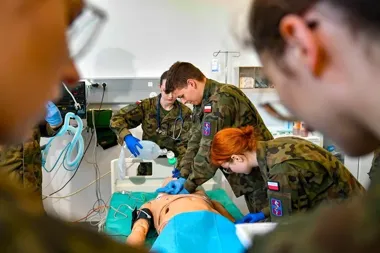 Żołnierze w mundurach moro ćwiczą medyczne zabiegi resuscytacyjne na fantomie w pomieszczeniu szpitalnym.