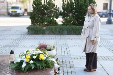 Kobieta w beżowym prochowcu stoi z opuszczoną głową przed kwiatami i zniczami na brukowanym placu miejskim.
