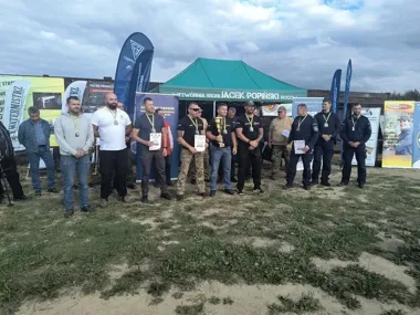 Grupa mężczyzn stoi na otwartym terenie przed namiotem i banerami podczas uroczystości wręczenia nagród po zawodach strzeleckich.