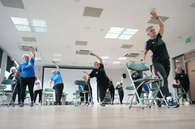 Grupa osób w sali gimnastycznej wykonuje ćwiczenia przy krzesłach podczas zajęć ruchowych.