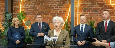 Kobieta w beżowej marynarce przemawia do mikrofonów podczas konferencji prasowej, za nią stoją cztery osoby na tle ceglanej ściany z neonami i roślinami.