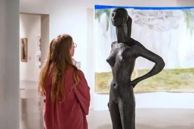 Kobieta ogląda stojącą rzeźbę kobiecej postaci w galerii sztuki, na ścianie za nią wisi obraz.
