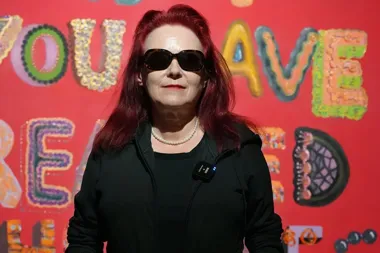 Kobieta z czerwonymi włosami w czarnej bluzie i okularach przeciwsłonecznych stoi przed czerwoną ścianą z kolorowymi, ozdobnymi literami.