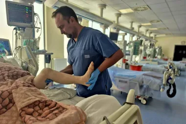 Mężczyzna w niebieskich fartuchach medycznych i rękawiczkach wykonuje ćwiczenia rehabilitacyjne nogę pacjenta na oddziale intensywnej terapii.