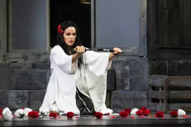 Kobieta w białym kimonie klęczy na scenie z mieczem przed sobą, otoczona czerwonymi i białymi różami, w tle kamienna sceneria teatralna.