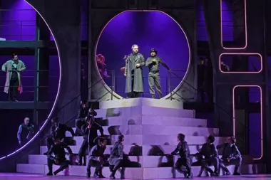 Aktorzy w mundurach i płaszczach na monumentalnej scenie z okrągłymi, podświetlanymi neonami podczas spektaklu teatralnego.