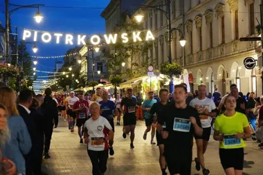Uczestnicy biegu ulicznego biegną wieczorem po ulicy Piotrkowskiej w Łodzi podświetloną neonowym napisem i lampkami.