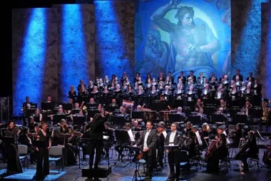 Orkiestra, chór i soliści występują na scenie podczas koncertu z monumentalną malowaną scenografią w tle.