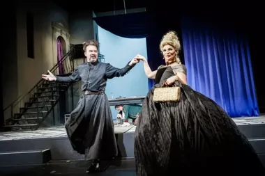 Aktor w kostiumie duchownego i aktorka w czarnej sukni z krynoliną tańczą na scenie teatralnej.