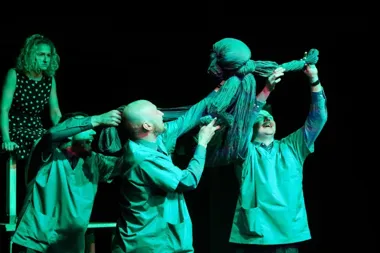 Aktorzy w zielonych fartuchach scenicznych manipulują dużą lalką wykonaną z materiału podczas spektaklu teatralnego.