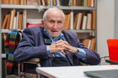 Starszy mężczyzna na wózku inwalidzkim siedzi przy stole w pomieszczeniu z regałami pełnymi dokumentów i książek.