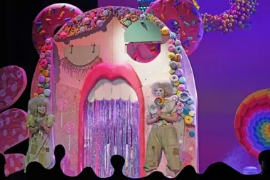 Dwie aktorki w kostiumach i perukach stoją przed kolorowym, słodkim scenograficznym domem z cukierkami i donutami podczas spektaklu teatralnego.