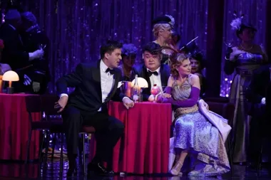 Trzy osoby w eleganckich strojach wieczorowych siedzą przy stoliku z czerwoną serwetą w teatralnej scenerii przypominającej kawiarnię lub klub nocny.
