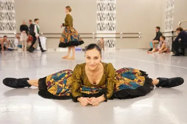 Tancerka w kolorowej, ludowej sukni leży w rozkroku na podłodze sali baletowej, w tle inni tancerzy i baletnicy przygotowują się do próby.