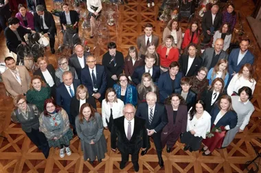 Grupa osób pozuje do pamiątkowego zdjęcia grupowego w reprezentacyjnej sali z drewnianą parkietową podłogą.