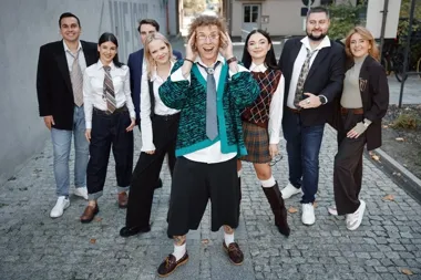 Grupa ośmiu osób w stylizowanych, szkolnych strojach pozuje na brukowanej uliczce, a mężczyzna na pierwszym planie trzyma się oburącz za głowę.