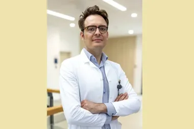 Mężczyzna w białym fartuchu lekarskim z założonymi rękami stoi w jasnym korytarzu szpitalnym.