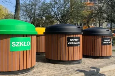 Cztery duże pojemniki do selektywnej zbiórki odpadów stoją na brukowanym placu w otoczeniu drzew i budynków.