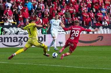 Piłkarz w czerwonym stroju o nazwisku Świderski strzela do bramki, a bramkarz w żółtym stroju o nazwisku Majchrowicz próbuje obronić podczas meczu na stadionie