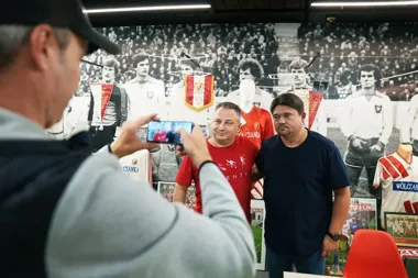 Mężczyzna robi telefonem zdjęcie dwóm mężczyznom stojącym przed wystawą pamiątek sportowych w pomieszczeniu muzealnym.