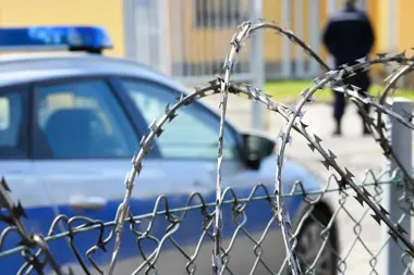 Policyjny radiowóz stoi za ogrodzeniem z drutem kolczastym, w tle widać sylwetkę funkcjonariusza.