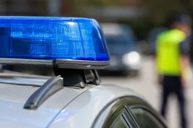 Błyskawica policyjna na dachu radiowozu, w tle rozmyty funkcjonariusz w kamizelce odblaskowej na ulicy.