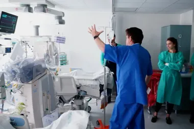 Personel medyczny w szpitalnej sali intensywnej terapii omawia przy łóżku pacjenta zabudowanym sprzętem monitorującym.
