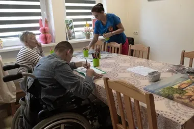 W jasnej sali przy stole z kredkami i kolorowankami mężczyzna na wózku inwalidzkim rysuje, starsza kobieta siedzi obok, a pracownica w niebieskim uniformie poda
